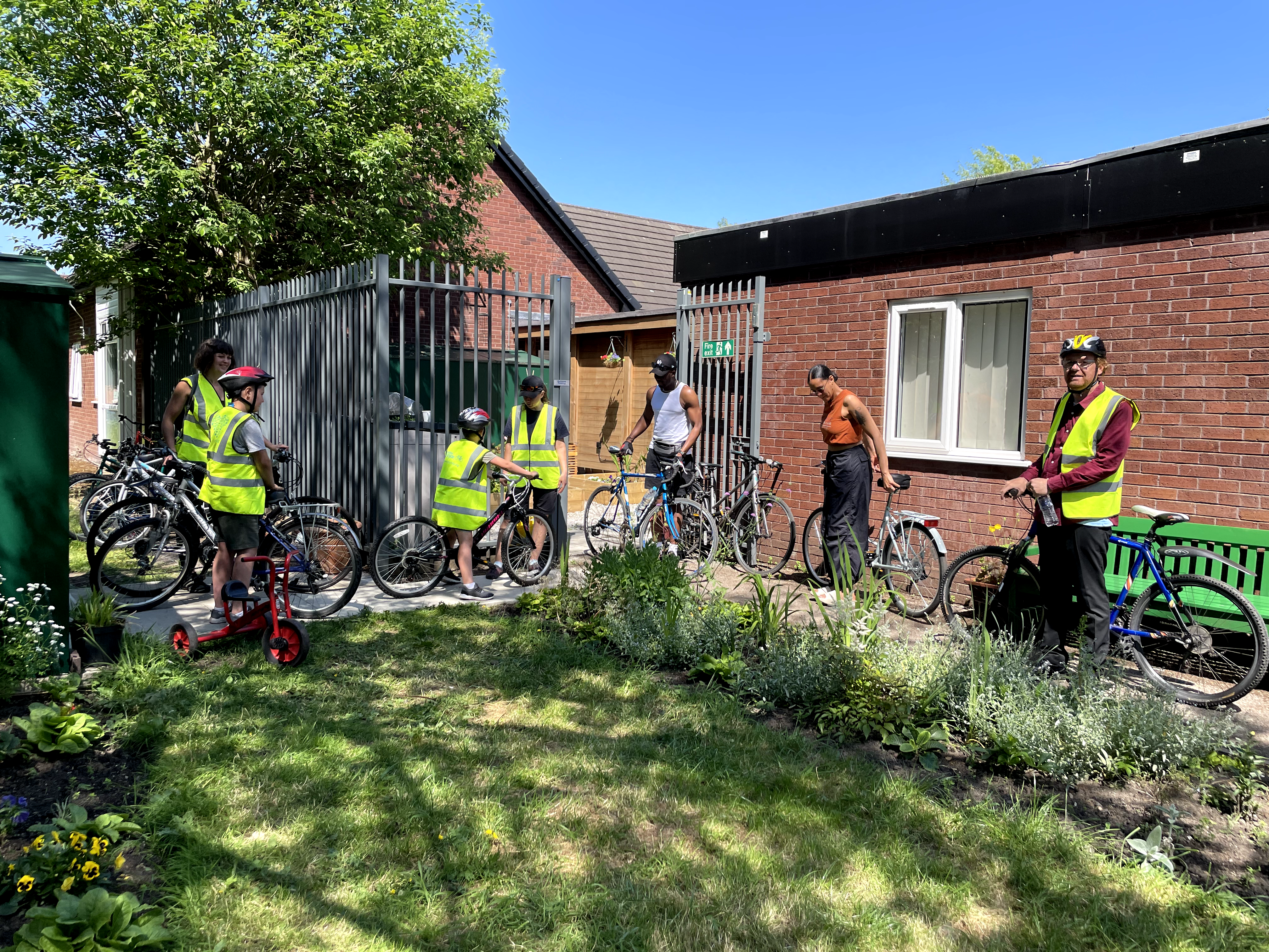 A group of people in high-visibility vests and helmets gathered outside a brick building with bicycles, preparing for a bike ride. One child is sitting on a bike, while another child stands near their bike. There is a small red scooter nearby. The scene is set in a garden area with green plants and trees on a sunny day.