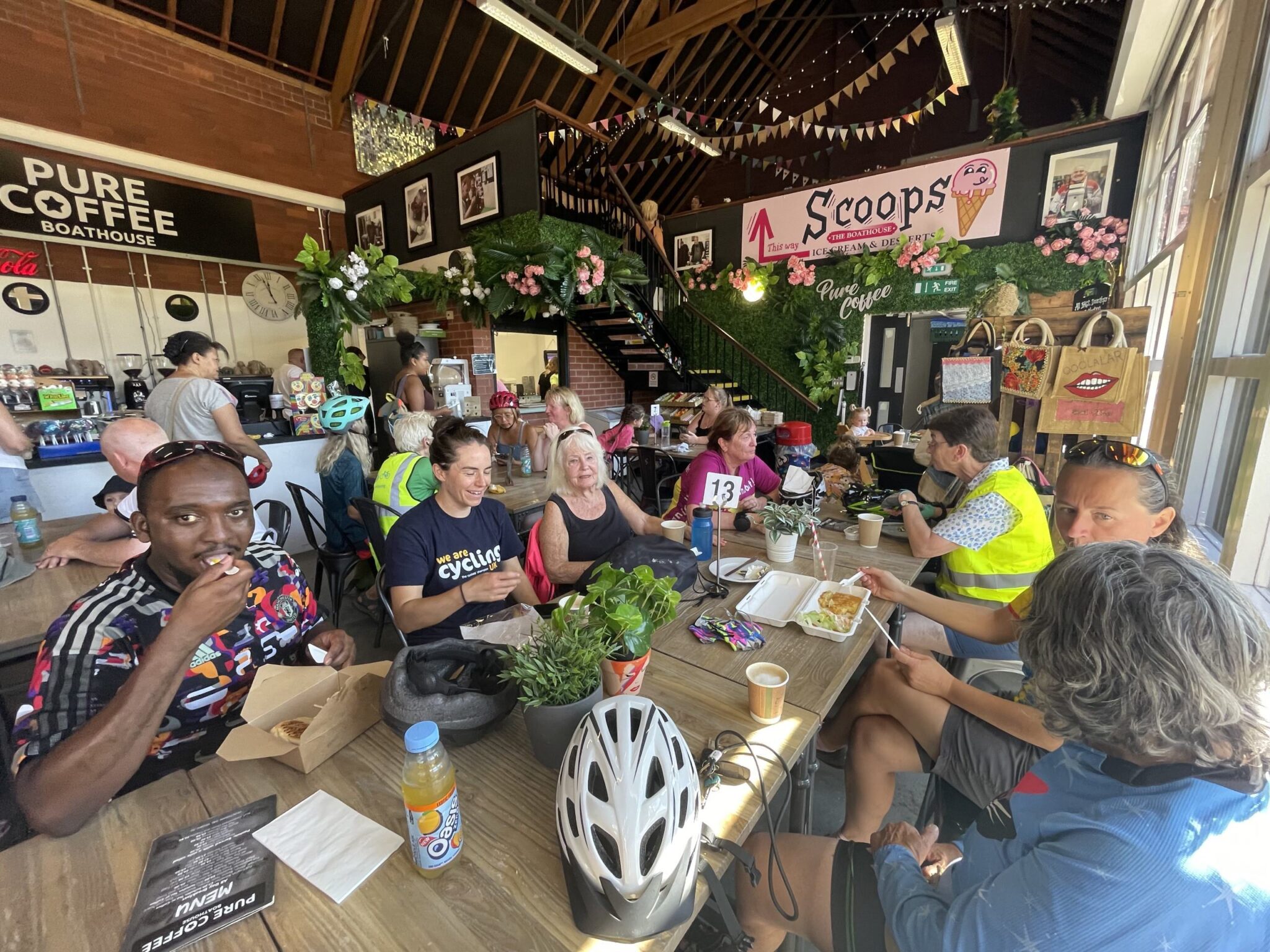 A group of people in cycling gear sit around a table in a café, eating and chatting, with bike helmets nearby. The café has decorative flowers and a sign that reads 'Pure Coffee' and 'Scoops'.